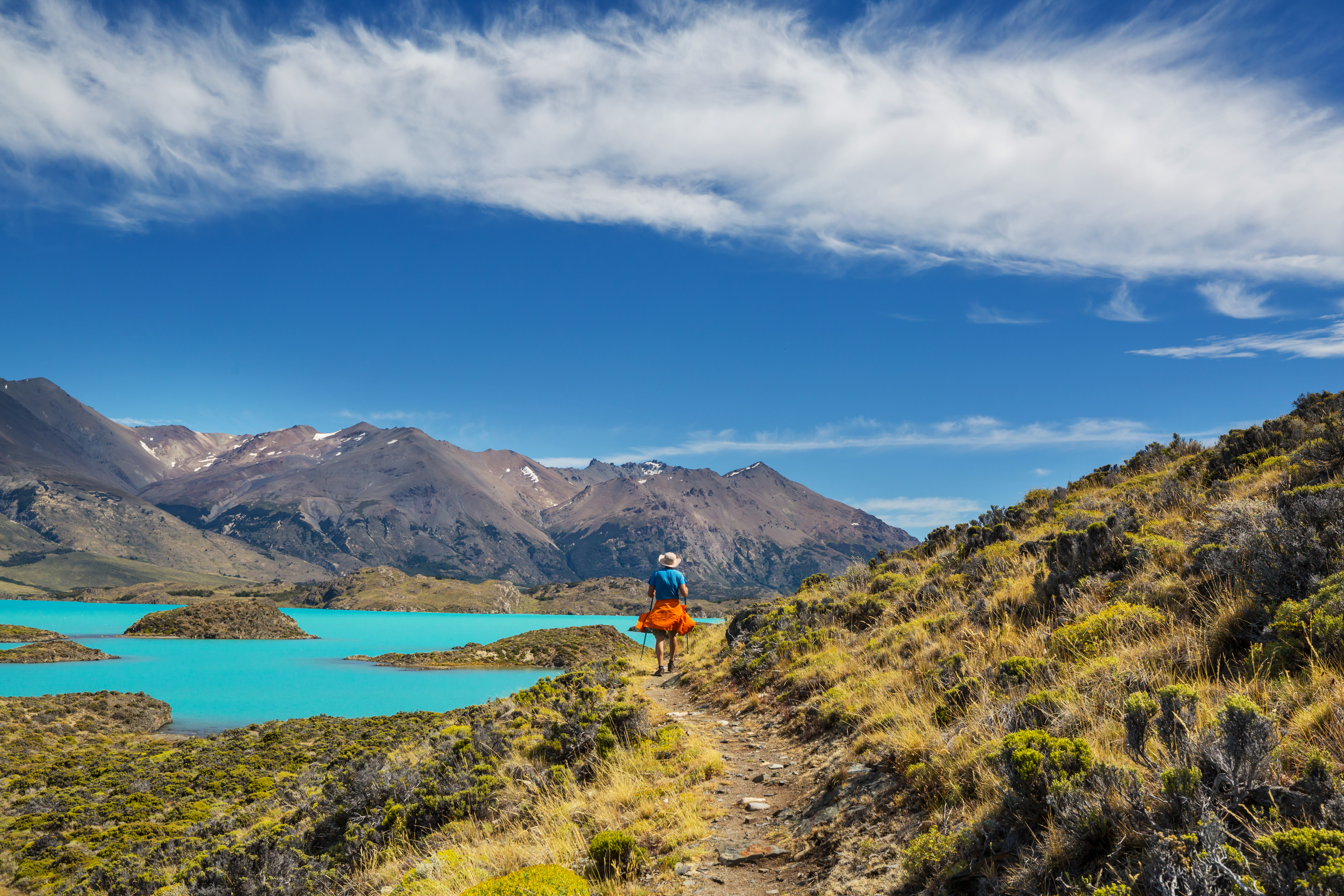 A traveler follows a narrow trail above a turquoise Patagonian lake toward the mountains