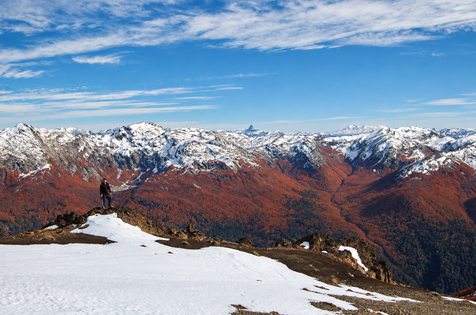 Jeff standing small on Cerro Colorado above autumn forests and a vast snowy mountain range near Villa La Angostura