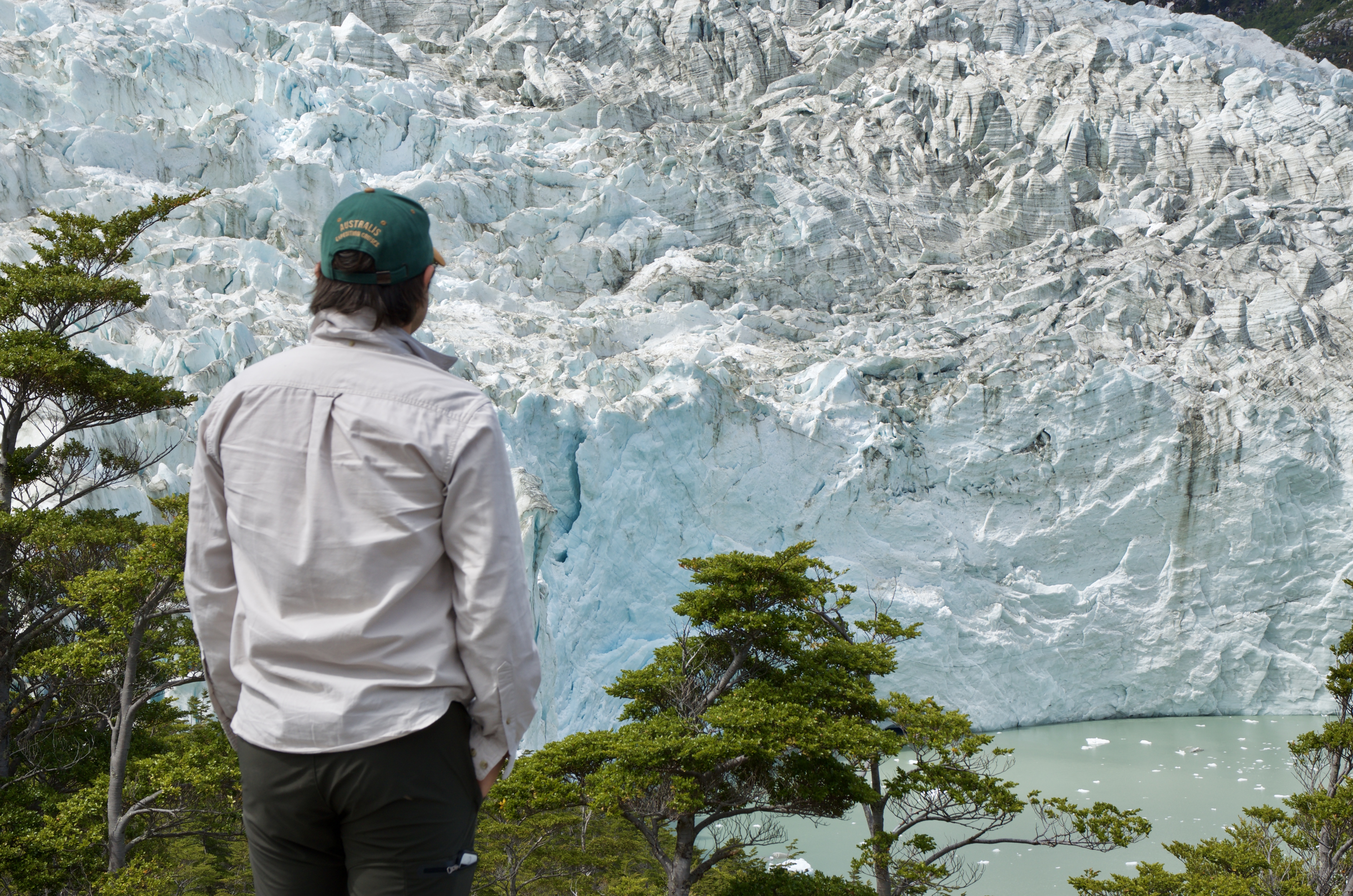 Jeff Gonzalez seen from behind, observing an immense glacier wall above the Patagonian forest and water
