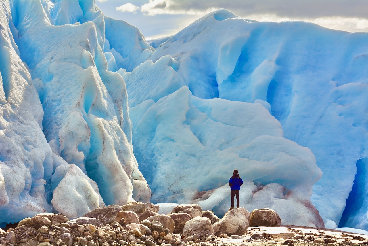 A traveler standing on rocks beside a towering wall of blue glacier ice